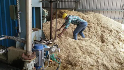Getty Images A worker, wearing turquoise-white checked shirt and blue jeans, inside a factory, working with stubble. 
