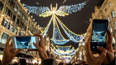 Two sets of hands holding up phones to take photos of the Angel shaped Christmas lights on Regent Street. White curved buildings line each side of the street.