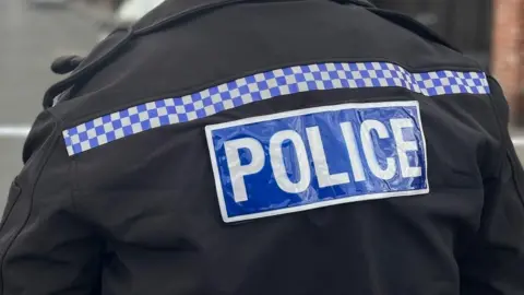 A stock image of a police officer pictured from behind. In the head-and-shoulders image he is wearing a black uniform jacket with the police logo on the back and a uniform hat. He is standing on a residential street where a police tape is cordoning off an area. The background is blurred.