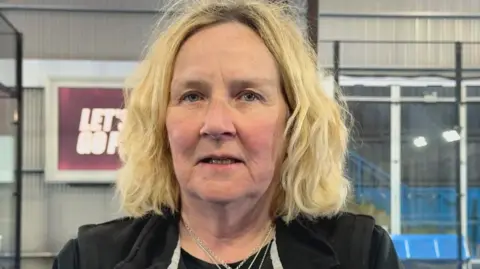 Jenny has medium blonde hair. She is wearing a black top. Behind her are two Padel courts, and a framed sign. She has a necklace around her neck. 