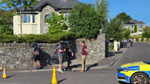 Councillor John O'Donoghue being interviewed by a TV crew in front of a wall surrounding the Homeland estate in Killarney. He has short dark hair and is wearing a red top and beige trousers. There is a blue and yellow garda car and yellow traffic cones in the foreground. Two officers in blue uniform are standing further into the estate, which consists of large detached houses. 