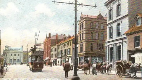 Courtesy of Peterborough Civic Society A tram in the middle of the street with horse-drawn carriages in front of shops and businesses.