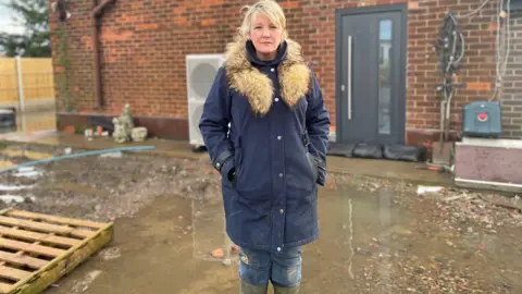 A woman with blonde hair stands in green wellies in flooded water.