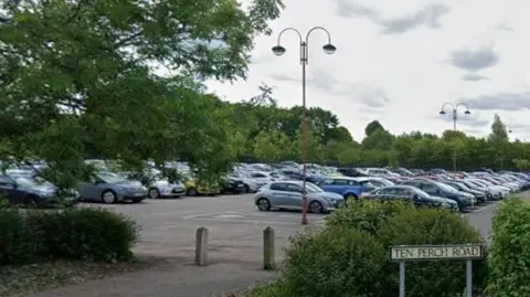 A park and ride car park filled with cars, A street sign in the foreground reads Ten Perch Road. 
