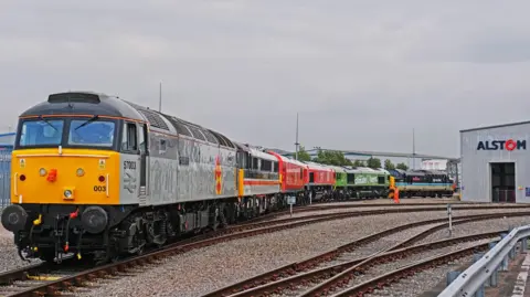 Ian Castledine A line of colourful locomotives rests on curved tracks outside an industrial building marked "Alstom"