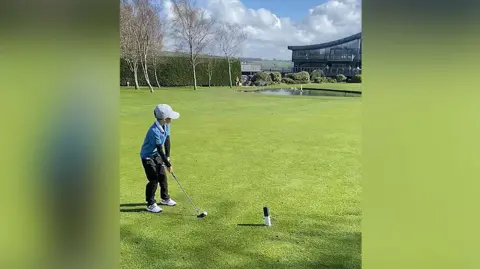 Official Hole in 1 Freddie wearing a light grey cap and blue gold top and black golf pants and white golf shoes tees off on Carus Green Golf Club course. 