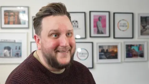 Lee Walker. He is looking past the camera while smiling. He is wearing a burgundy jumper and standing in front of a wall on which there are various framed images.