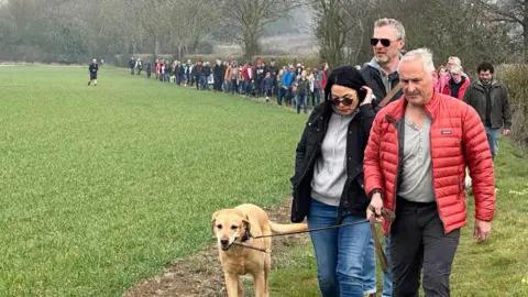 Jenny Kirk/BBC Dozens of people walking around the outside of a field, with a man in a red jacket, grey top and black jeans at the front, with a golden labrador on a lead. A woman beside him has dark hair, black sunglasses, a black jacket, grey top and blue jeans