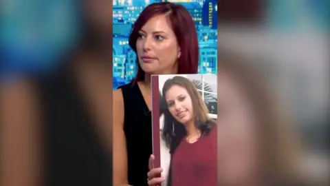 A woman with shoulder length brown hair wears a black vest top. She is holding a blown up photo of her younger self.