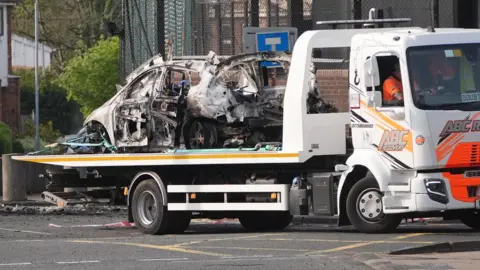 Niall Carson/PA The remains of an exploded vehicle are removed from the scene outside the police station in Dunmurry. The vehicle was hijacked shortly after 10.50pm on Saturday in the Twinbrook area of west Belfast and a gas cylinder device was placed in the boot. Homes in the area, which is on the outskirts of Belfast, have been evacuated and members of the public have been advised to avoid the scene. Picture date: Sunday April 26, 2026. PA Photo. Photo credit should read: Niall Carson/PA Wire
