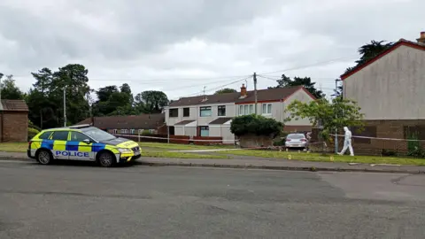 A police car parked at the side of the road in front of a row of houses. There is a forensics police officer walking along the verge and police tape.
