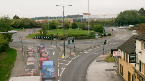 A large roundabout is shown with multiple roads leading into it.
Several traffic lights around the junction are illuminated.
A line of vehicles waits at a red light in the foreground.
Trees and bushes surround the roundabout, with buildings visible in the background.