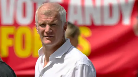 Al Carns outside on a very sunny day in a white shirt against a red banner looking at the camera.