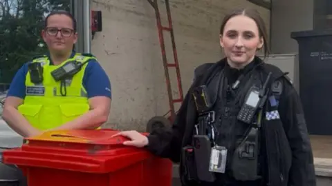 Two police officers are standing next to a red knife amnesty bin. One of the police officers is wearing a high visibility jacket with police radio and body camera attached. The other officer is wearing a black police uniform and also has a police radio and body worn camera attached to her jacket.