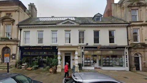 Google Two-storey building with tall windows on the top floor and a dormer window in the roof. The first floor is rendered in white, while the ground floor has a dark-coloured plant shop and a lighter-coloured estate agents shop. Between the two shops is a pillared entrance way in white and beige, with a small porch leading to a dark wood door.