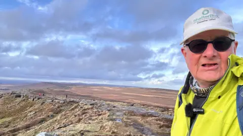 ShelterBox Dave Foster talking a selfie with a vast moorland in the background. He is wearing a white cap with ShelterBox written on it, black sunglasses, and a high-vis waterproof jacket.