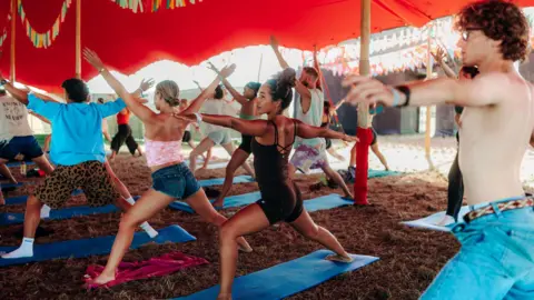 Boardmasters Festival / Josh Harkon a class of people practicing yoga on mats under a tent in the sunshine. 
