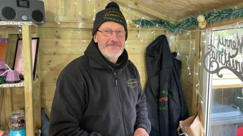 A man in a black jumper with black beanie in a wooden shed with Christmas decorations