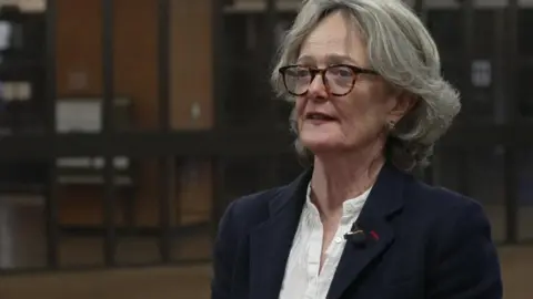 Elizabeth Campbell, a woman with short wavy hair and tortoiseshell glasses. She wears a navy jacket over a white collarless blouse.