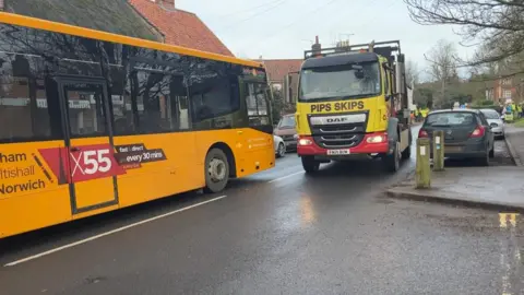 Andrew Turner/BBC A yellow bus marked the X55, and a yellow and red jump truck faces in the B1150 in Coltishall. There are cars parked on both sides of the road, and an incorporation of pavement on the road to make a pedestrian crossing point.