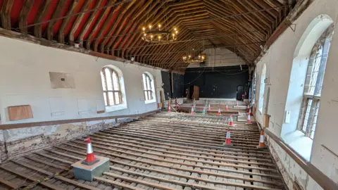 Borough Council of Kings Lynn & West Norfolk Inside the guildhall's roof. There are wooden beams over the floor and orange traffic cones on top of these. The roof is arched with lots of large wooden beams. 