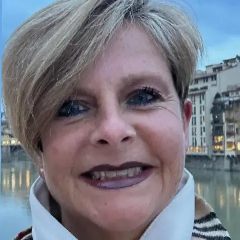 Jane Rowlands-Mellor Jane Rowlands-Mellor is wearing a striped scarf and is stood in front of a river with buildings in the background.