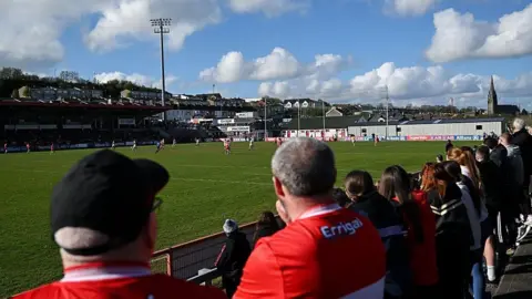 Getty GAA players are playing on a large grassy pitch and a number of people in Derry GAA shirts are pictured watching at the sidelines. A large floodlight can be seen towering above the stadium and the city skyline is seen in the distance. 