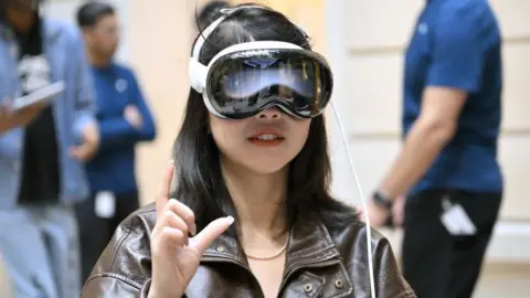 Getty Images Uma mulher usando o headset Vision Pro dentro de uma loja da Apple. Parece um par de óculos elegante, brilhante, curvilíneo e futurista preso ao rosto dela. Você quase diria que parece legal.