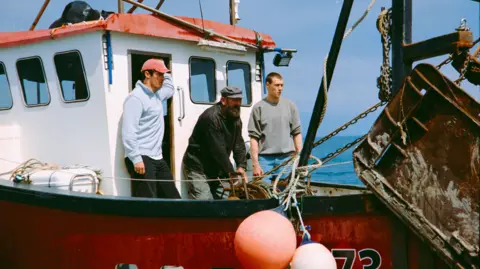 Steve Tanner/Bosena The image shows three men standing on the deck of a small fishing boat. It is a sunny day and the sea is calm. One of the men appears to be winching some up with a metal chain.