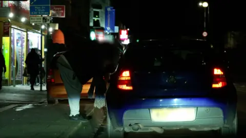 A blue car pulled up to the curb on a street at night. A woman is talking to the people in the car. Her face is blurred and it is dark, so she is hard to make out.