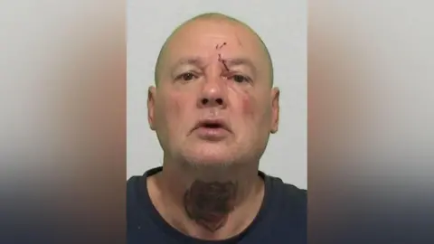 Mugshot of Straughan. He is bald with a cut above his left eye and a large red rose tattooed across the front of his neck
