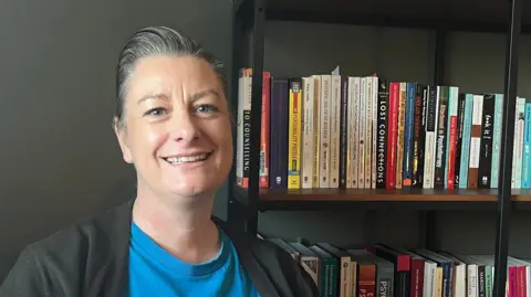 Zoe Hughes, an Exeter City council member, wearing a blue T-shirt and a black top, sitting next to a bookshelf