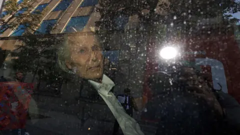 AFP via Getty Images Peter Nygard is photographed through a car window. He looks towards the camera, and has long white hair, and wears a black suit jacket and white button-up shirt.
