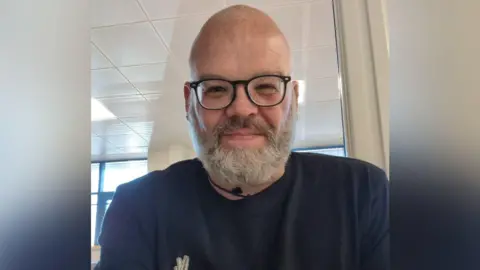 A selfie of a bearded and bespectacled man who smiles at the camera. He is wearing a blue top with a white logo on his chest.
