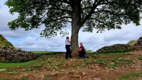 Hayley Graham-Hardy Hayley Graham-Hardy is standing side on with her hands to her face wearing black trousers and a white top below the Sycamore Gap tree which stands with Hadrian's Wall behind. Lee Graham-Hardy is on one knee proposing to her. He's wearing a back top and trouser.