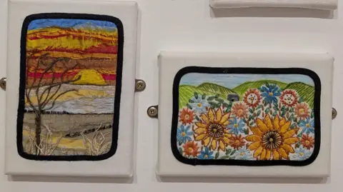 Darlington Stitch and Create Two pieces of embroidery have been placed on a white wall. One shows a sunset with a bare tree in the foreground and the other shows two sunflowers with green fields in the background.
