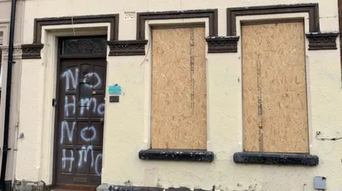 A beige terraced house with boarded up windows and graffiti on the door.