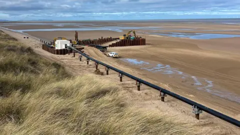 Eni This image shows Talacre Beach, There's construction works happening in the background, The long line of structures now have a black pipe resting on top of them. There's long grass and sand around the construction site.