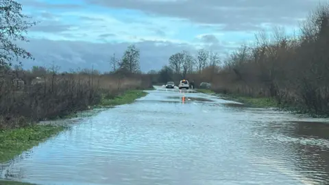Scott Caseling A flooded road in Somerset. There are two vehicles in the distance. One is attempting to drive through flood water.