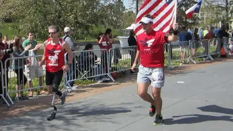 Chris and Denise Arthey Two runners on a road running past spectators. One runner has a prosthetic leg and the other runner is carrying an American flag. 