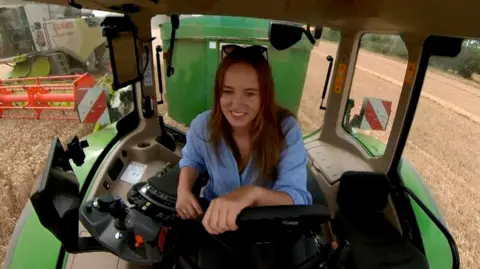 Steve Hubbard/BBC Alice Northern smiling while driving a tractor. The camera is inside the cab and we are looking at Alice as she steers the tractor, which has pulled up next to a combine harvester. One of her hands is resting on the tractor's wheel and she is sitting a little forward in the seat. She is wearing a light blue shirt and has a pair of sunglasses on her head.