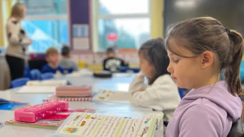 A classroom setting with several children seated at a table, working on colorful activity books. The table has open books, a pink pencil case, and a green water bottle. In the background, a teacher stands near a cabinet, and a large digital screen is mounted on the wall. The classroom is bright with large windows and colorful decorations.