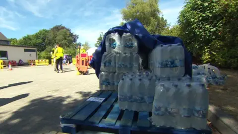 Multiple cases of bottles water sit on a pallet in a paved outdoor space. Some people in hi-vi jackets are in the background.