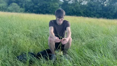Aidan is crouched down in a green field. He has brown hair and wears glasses and is playing with a few blades of grass. He's wearing a brown T-Shirt and shorts.