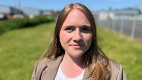 Laura young with long auburn hair. She is standing on grass and wearing a brown jacket over a white top.
