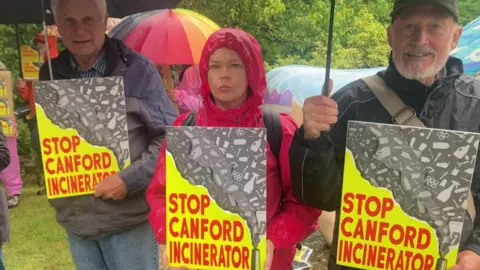 Three protesters, under umbrellas, holding yellow and red placards with the words 'STOP CANFORD INCINERATOR'