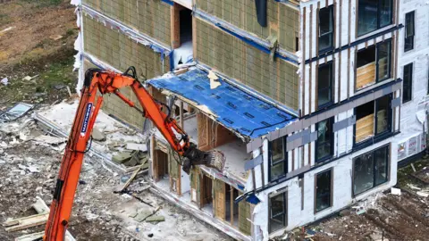 Eddie Mitchell An orange digger tearing chunks from a block of flats