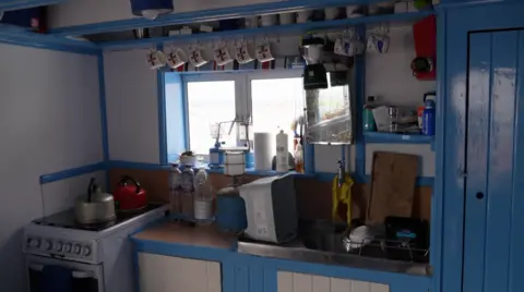 Inside Richard Miles' hut, a small, functional kitchen with white walls and bright blue trim and cabinetry. A row of mugs hangs from hooks above a window, which looks out onto a coastal view. On the left, a white stove holds two kettles, while the countertop contains water bottles, a gas canister, and various kitchen items. A stainless-steel sink sits beneath shelves stocked with bottles, cups, and supplies. The space is neatly arranged and brightly painted, with natural light entering through the central window and reflecting off the blue woodwork.