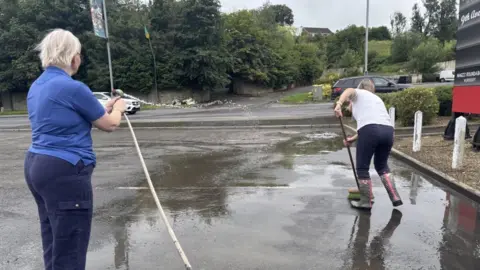 A person dressed in blue holds a hose on a forecourt at Sligo Road in Enniskillen, while another person in white top and blue trousers sweeps up.