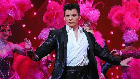 Getty Images Nigel Harman in I Can't Sing: The X Factor Musical. He is dressed to represent Simon Cowell with high-wasted black shiny trousers and jacket and a white shirt and braces. The shirt is open to the chest. He has short black hair and is dancing. Behind him are dancers in bright pink sequinned costimes with feather head dresses and pink lighting.